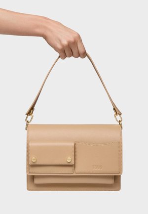 Mano sosteniendo un bolso de cuero beige con dos bolsillos frontales, herrajes dorados y correa corta contra un fondo blanco liso.