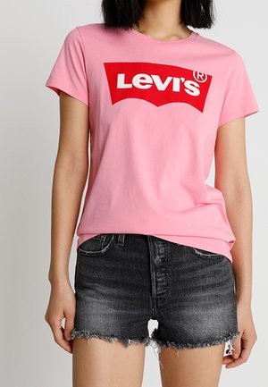 Kvinde iført en lyserød Levi's-logo t-shirt og sorte denimshorts med frynser, stående mod en ensfarvet lys baggrund.