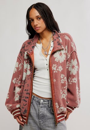 Le mannequin porte une veste zippée rose à motif floral par-dessus un haut blanc avec des colliers superposés et un jean gris sur un fond de studio uni.