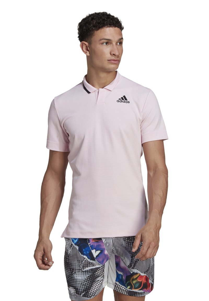 Polo shirt rose clair avec un design texturé, col côtelé et logo Adidas noir sur la poitrine gauche. Assorti à un short à motifs colorés.