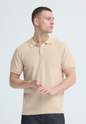 Man met kort haar, gekleed in een beige textuur korte mouwen polo, die zijn handen samenhoudt en naar rechts kijkt tegen een effen lichte achtergrond.