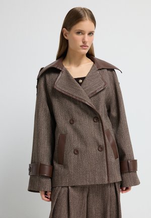 Bruine tweedjas met bruine leren accenten, dubbelgenoemde ontwerp, brede kraag en grote knopen. Heeft twee zijzakken.