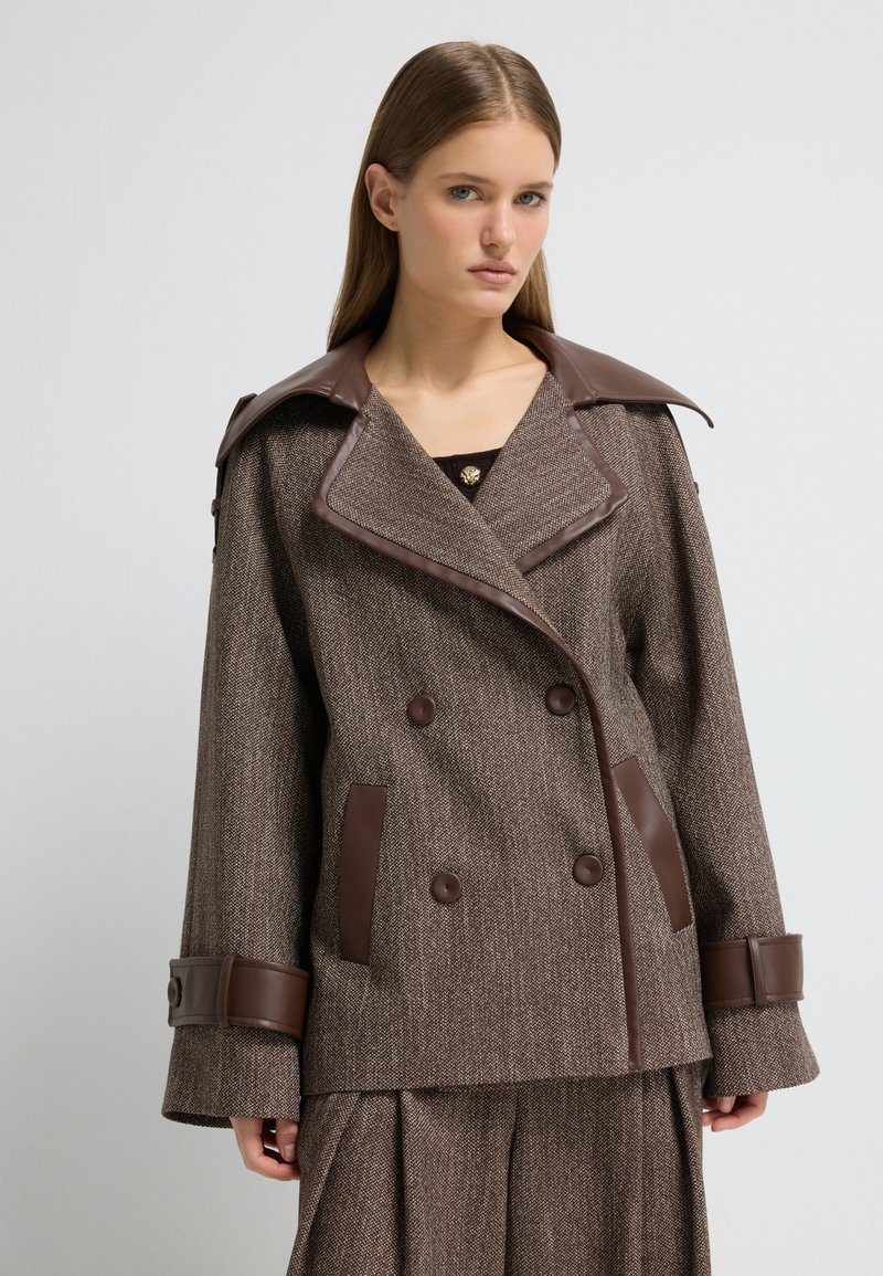 Giacca in tweed marrone con dettagli in pelle marrone, design doppiopetto, ampio colletto e grandi bottoni. Presenta due tasche laterali.