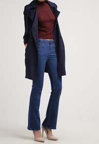 Marinblå trenchcoat över en vinröd ribbad tröja, matchad med utsvängda blåjeans och beige högklackade skor. Släta texturer och skräddarsydd passform.