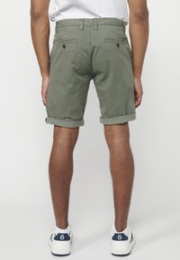 Muškarac u maslinastozelenim šortsevima podvrnutim do koljena i bijelim tenisicama, prikazan s leđa na jednostavnoj bijeloj pozadini.