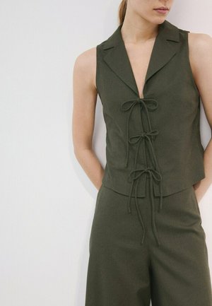 Femme portant un gilet sans manches vert foncé avec trois fermetures à nouer à l'avant et un pantalon taille haute assorti sur un fond blanc uni.