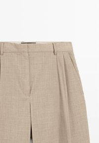Pantalons beiges ajustés avec pince, fermeture éclaire sur le devant, passants de ceinture et poches latérales sur fond blanc.
