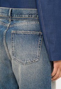 Modra denim hlače s svetlim pranjem, ki imajo zadnji žep in visok pas. Delno je vidna modra jakna s kontrastnim šivanjem.