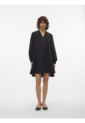 Femme portant une robe noire à manches longues boutonnée avec un col en V et des chaussures slingback noires à bout pointu, se tenant devant un fond blanc uni.