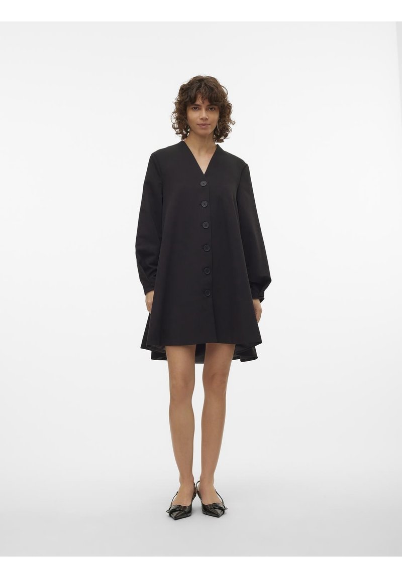 Femme portant une robe noire à manches longues boutonnée avec un col en V et des chaussures slingback noires à bout pointu, se tenant devant un fond blanc uni.