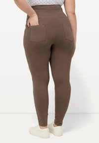 Leggings marroni a vita alta con due tasche posteriori, texture morbida e silhouette aderente, abbinati a sneakers bianche.