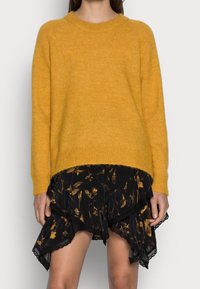 Frau in senfgelbem Strickpulli und gestuftem schwarzem Rock mit Blumenmuster und Spitzenbesatz, die vor einem schlichten hellen Hintergrund steht.