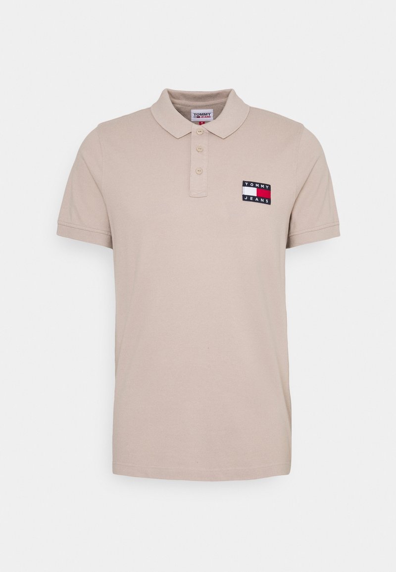 Beige pikétröja i bomull med krage, tre knappar och en färgglad logotyp på vänster bröst. Korta ärmar.