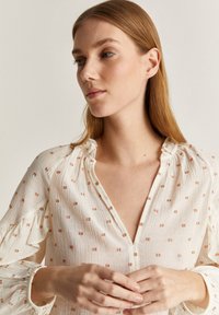 Blusa leggera bianca con scollo a V e colletto arricciato. Presenta piccoli motivi quadrati color rame su tutta la superficie e maniche ampie.