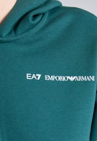 Τιρκουάζ φούτερ φτιαγμένο από μαλακό ύφασμα με ριγέ υφή. Διαθέτει το λευκό λογότυπο "EA7 EMPORIO ARMANI" εκτυπωμένο στο μπροστινό μέρος.