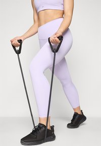 Leggings attivi color lavanda chiaro abbinati a un reggiseno sportivo coordinato, mentre si impugnano bande di resistenza nere e si indossano sneaker sportive nere.