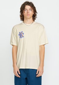 Camiseta de algodón beige con corte relajado, que presenta un gráfico geométrico azul y naranja en el área del pecho izquierdo. Mangas cortas y cuello redondo.