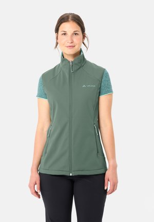 Frau trägt eine grüne ärmellose Weste mit Reißverschluss über einem türkisfarbenen Kurzarmshirt und schwarzen Hosen, steht vor einem schlichten weißen Hintergrund.