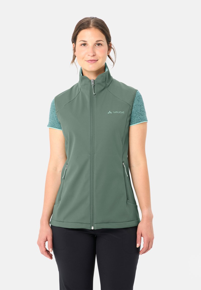 Frau trägt eine grüne ärmellose Weste mit Reißverschluss über einem türkisfarbenen Kurzarmshirt und schwarzen Hosen, steht vor einem schlichten weißen Hintergrund.