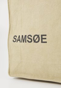 Beige canvas tote bag featuring the printed brand name "SAMSØE" in bold black letters. The fabric has a textured appearance.