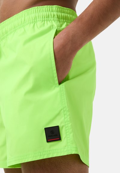 Neonově zelené plavky s elastickým pasem, boční kapsou a černým logem na levé noze. Hladká, lehká tkanina.