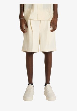 Mężczyzna w kremowych plisowanych szortach, dopasowanej koszuli zapinanej na guziki, białych trampkach i czarnych skarpetkach, stojący na jednolitym tle.