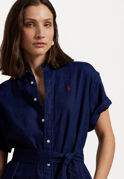 Jūras zila, īsām piedurknēm krekldrēbju kleita no viegla auduma ar jostu viduklī, ar smalku sarkanu logo uz krūtīm un pogām.