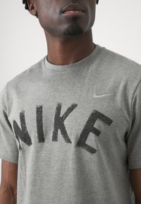 Pelēka kokvilnas T-kreklu ar lielu melnu "NIKE" teksta dizainu. Ietver mazu pelēku Nike swoosh kreisajā augšējā krūšu rajonā. Īsas piedurknes.