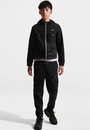 Chłopiec stojący, ubrany w czarną bluzę Nike na suwak z kieszeniami, dopasowane czarne spodnie bojówki i czarne buty sportowe na białym tle.