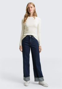 Jonge vrouw met lang blond haar, gekleed in een nauwsluitende witte top met lange mouwen, donker wijde broek met opgerolde pijpen en witte sneakers, staand rechtop.