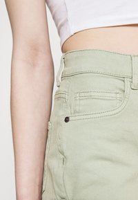 Vihreät denimpantalonit, joissa on tekstuuripinta. Ominaisuuksiin kuuluvat vyötärönauhan naru, vyölenkit ja etutasku metallitulppineen.