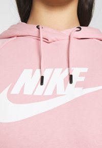 Detailní záběr na osobu v růžové mikině s kapucí, na které je bílé logo Nike a šňůrky s černými konci kolem krku.