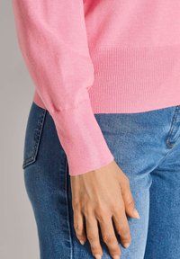 Helles rosa gestricktes Sweatshirt mit gerippten Bündchen, kombiniert mit blauen Denim-Jeans. Nahaufnahme des Ärmels und der Hand, die auf dem Oberschenkel ruht.