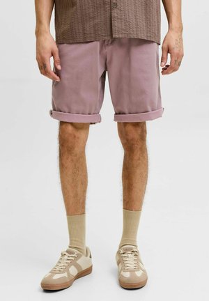 Man wearing mauve rolled-up shorts, beige crew socks, and beige casual sneakers, standing with hands relaxed by sides.