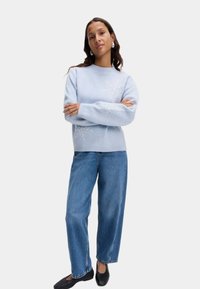 Jasnoniebieski sweter z haftem w kształcie płatków śniegu, ze ściągaczem przy dekolcie i mankietach. Połączony z niebieskimi spodniami damskimi z wysokim stanem i czarnymi balerinami, stoi z założonymi na piersi rękami.