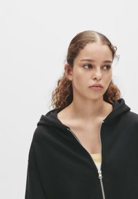 Zwarte zip-up hoodie met een relaxte pasvorm en een capuchon. De stof lijkt zacht, met een gladde textuur. Een lichtgele top is zichtbaar eronder.