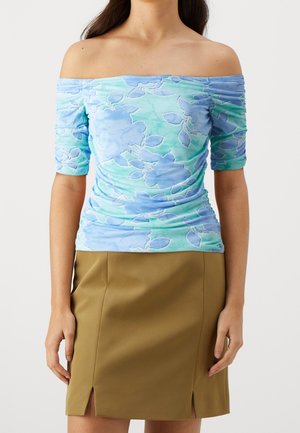 Vrouw draagt een geplooide off-shoulder top met blauwgroene bloemenpatronen en een knielange bruine rok met voorsplitten.