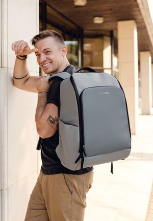 Lächelnder Mann mit kurzen Haaren und Tätowierungen, der ein schwarzes T-Shirt und beigefarbene Hosen trägt, steht neben einem Gebäude und trägt einen großen grauen Rucksack.