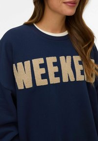 Femme portant un sweat-shirt bleu marine avec des lettres beige en relief formant le mot "WEEKEND" sur la poitrine et un sous-pull blanc à col rond.