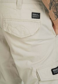 Lichtbeige cargoshorts van geweven stof met een zak voorzien van een zwart label. De textuur lijkt glad met een lichte glans.