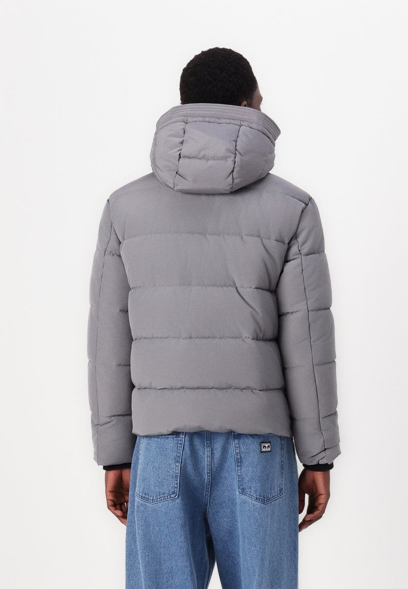 Chaqueta acolchada gris con capucha, costuras horizontales y un corte relajado, combinada con pantalones de mezclilla azul.