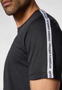 Μαύρο αθλητικό μπλουζάκι με κοντά μανίκια, με μια λευκή κάθετη ρίγα λογότυπου κατά μήκος του ώμου. Λείο ύφασμα, σχέδιο με λαιμόκοψη.