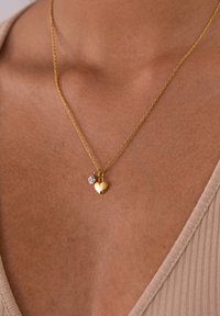 Gold-Halskette mit einer verdrehten Kette, versehen mit einem goldenen Herzanhänger und einem kleinen klaren Edelstein. Schmuck liegt auf einem hellen Hautton.