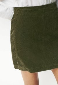 Fustă din catifea cord cu un verde măsliniu, cu nervuri verticale, croi drept și hem care se oprește deasupra genunchiului. Prezintă o textură și o structură subtile.