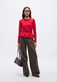 Rød langermet topp med innsvinget midje og rysjekant på kanten, kombinert med vide brune bukser. En liten brun håndveske fullfører antrekket.