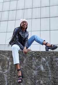 Svart läderjacka, grafisk t-shirt, ljusblå jeans, svarta plattformsskor med ankelremmar, bärande en ljusrosa mössa, sittande på en stenmur.