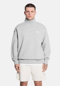 Mężczyzna stoi twarzą do kamery, ubrany w jasnoszary sweter na zamek i białe szorty, z widocznymi tatuażami na ramionach.