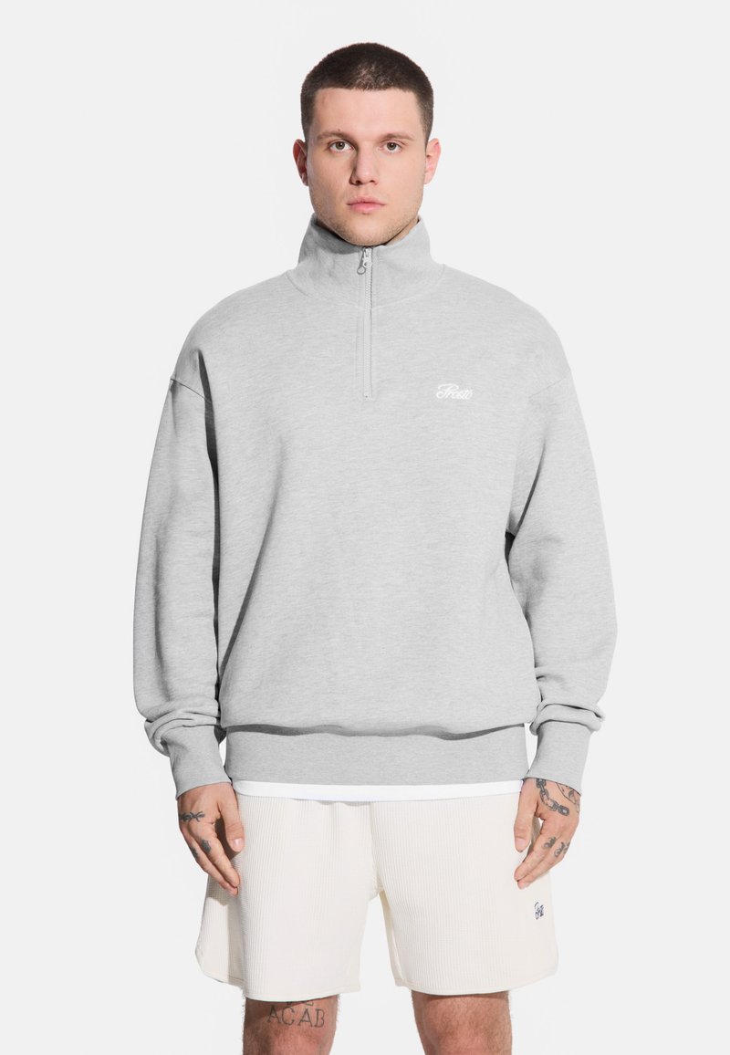 Mężczyzna stoi twarzą do kamery, ubrany w jasnoszary sweter na zamek i białe szorty, z widocznymi tatuażami na ramionach.