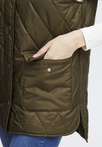 Gestepptes olivgrünes Weste mit einer Seitentasche mit Druckknopfverschluss und einem Schlitz am Saum. Das Material hat eine glatte, matte Textur.