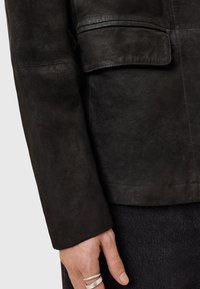 Chaqueta de ante negra con un diseño elegante, con un bolsillo y detalles de costura, complementada por un acabado texturizado. Mano que muestra anillos de plata.